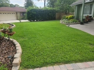 Well-maintained backyard with lush green lawn, stone border edging, landscaped flower beds, and house structure visible in background